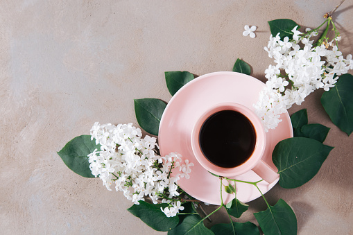 High angle view of black coffee in pastel pink cup surrounded with green leaves and white lilac flowers on rustic brown stone background with copy space.