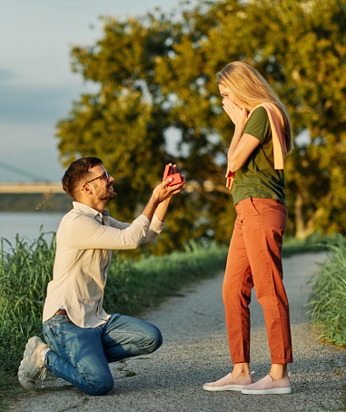 Cheerful handsome man proposing to his girlfriend. Portrait of a young happy couple having fun outdoors in nature during sunset. Girlfriend and boyfriend bonding, love concept during summer.