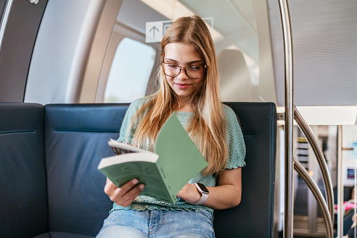 Beautiful teenage girl sitting in a modern fast train and reading a book. Public transport concept.