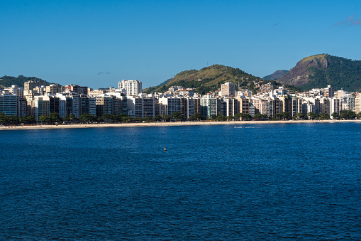 View of the edge of Praia de Icaraí, a neighborhood in Niterói, Rio de Janeiro, Brazil. Buildings and houses in front of the beach, surrounded by hills and nature. Bathed by Guanabara Bay. Sunny day.