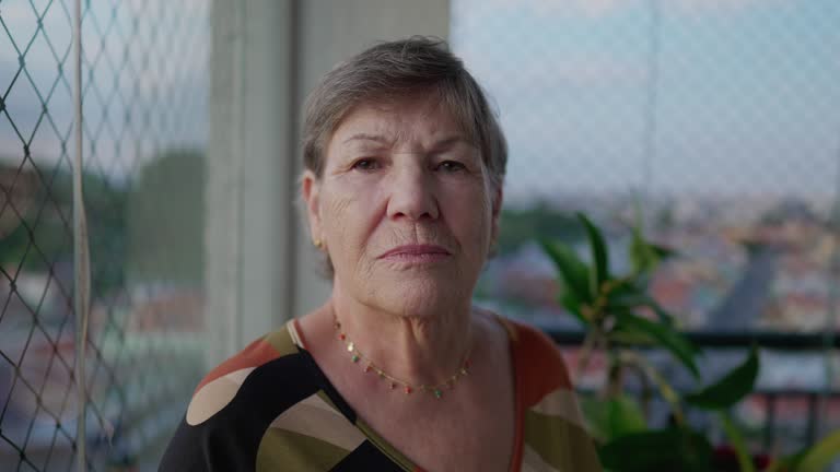 Portrait of a serious elderly woman face looking at camera standing at apartment balcony looking at camera