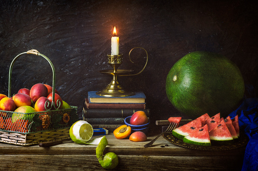 Classic still life with various fruits placed in vintage basket, watermelon slices, old books and illuminated candle on rustic wooden background.