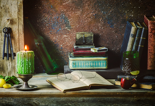 Classic still life with vintage books placed with illuminated candle, old boxes, pipe, antique keys, lemon, mint and drink on rustic wooden background.