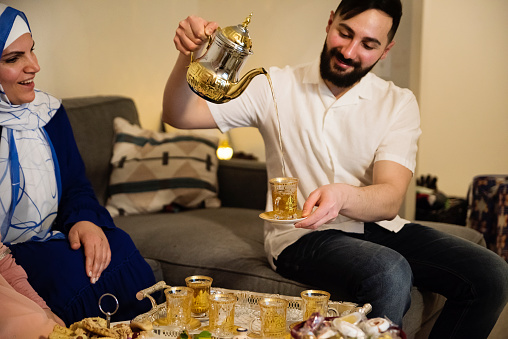 North-American muslim family eating Iftar at Ramadan. Traditional Lebanese and moroccan desserts on the table. Father is serving tea, mother is wearing a hijab. Horizontal waist up indoors shot. This was taken in Quebec, Canada.