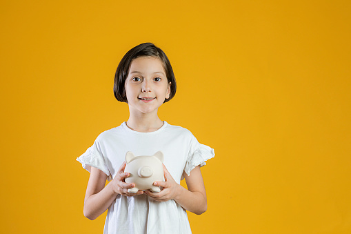 Little girl in white dress looking at camera holding her piggy bank  in front of yellow background. Pocket money and savings concept.