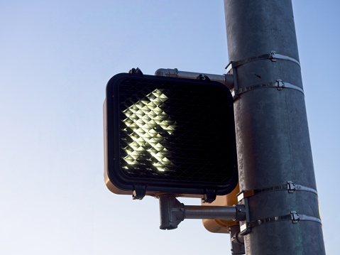 A pedestrian walk sign light against a cold blue sky with copy space. Crosswalk light on metal pole near busy intersection.