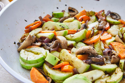 Delicious homemade spring vegan meal with fresh vegetables. carrot, mushrooms, zucchini, in a cooking pan, with seasoning and herbs on a white rustic wooden kitchen or restaurant table, representing a healthy lifestyle, wellbeing and body care, top view with a copy space