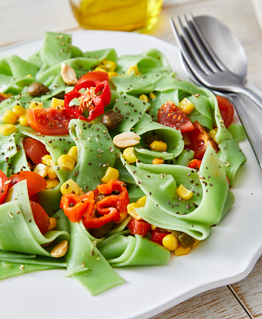Healthy green Vegan pasta, made of spinach, corn, red pepper and garlic, served on an elegant ceramic plate, with fresh vegetables and herbs around, lemon, olive oil and pepper, on a white rustic wooden natural kitchen or restaurant table, representing a wellbeing and a healthy lifestyle and food indulgence, top view image with a copy space