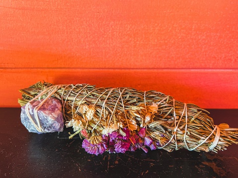 Horizontal closeup photo of dried, scented sweet smelling grasses and Rosemary leaves, an Amethyst crystal and a small bunch of tiny coloured dried Statice flowers wrapped with white cotton string, to make a Smudge Stick, the smoke of which is used to purify spaces, on a wooden bench in front of a vibrant orange painted wooden wall, on display in a cafe, Byron Bay, NSW.