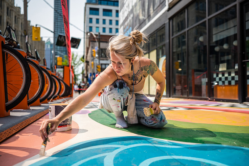 Young Caucasian woman artist painting sidewalk mural. She is dressed in casual work clothes. Exterior of public sidewalk in downtown of large North American City.