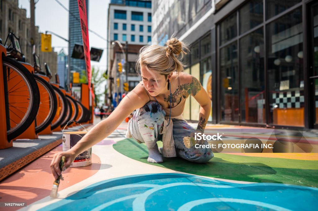 Young Caucasian woman artist painting sidewalk mural Young Caucasian woman artist painting sidewalk mural. She is dressed in casual work clothes. Exterior of public sidewalk in downtown of large North American City. Painting - Activity Stock Photo