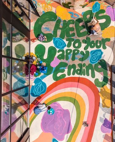 Two Female artists painting large wall mural. They are dressed in casual work clothes. Exterior of public  sidewalk in downtown of large North American City. Drone point of view.