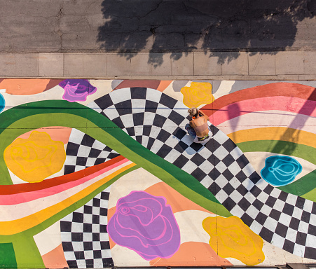 Young Caucasian woman artist painting sidewalk mural. She is dressed in casual work clothes. Exterior of public sidewalk in downtown of large North American City. Drone point of view.