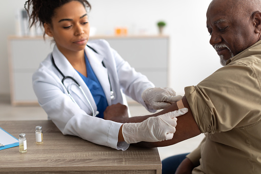 Close up of black female doctor in protective gloves applying sticking patch to patient's arm after injecting elderly man with seasonal flu or infection vaccine, selective focus on hand