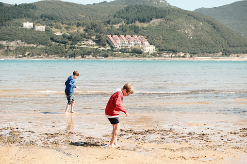 Siblings playing on the beach. It's springtime. It's still cold. They play at getting their feet wet. Dressed in tracksuits and coats. Long pants rolled up. Cold water. Atlantic Ocean. Mountainous coast. Sand with seaweed.