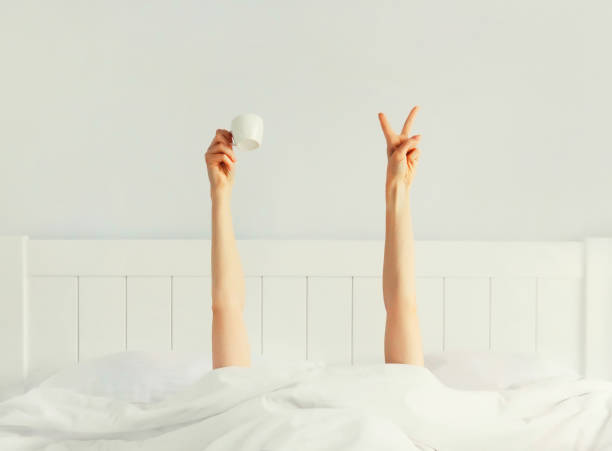 istock Cheerful lazy woman waking up after sleeping lying in soft comfortable bed showing empty cup coffee stretching her hands up from under the blanket in white bedroom 1492732193
