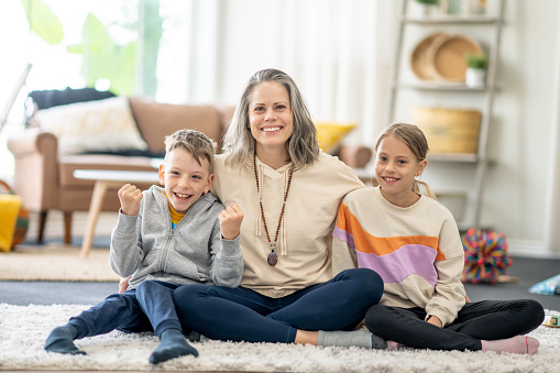 A Mother sits with her two kids on the floor in the comfort of her own living room as they pose for a portrait.  They are each dressed casually  and are smiling.