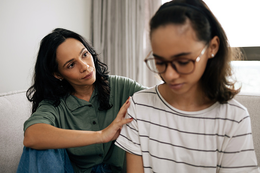 Caring mother comforting her teenage daughter, both sitting on the couch at home, sharing a tender and supportive moment