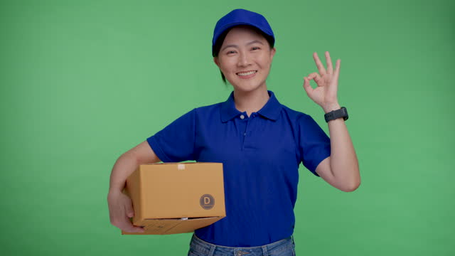 Asian woman in uniform polo t-shirt holding box happy smiling with confident showing okay sign standing isolated over green background.