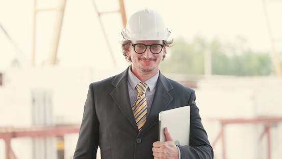 Professional investor or businessman or real estate business owner have leadership and vision, standing confident at construction site of real estate projects. Male construction manager in suit with helmet on head standing and holding laptop and looking at camera.