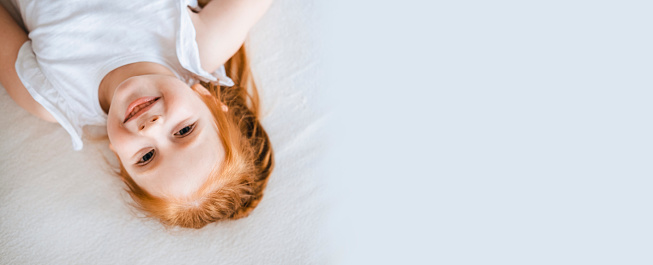 Red-haired girl lies on white bed linen, top view. Charming child with bright hair and freckles. Banner with place for text.