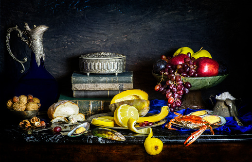 Classic still life with fresh fruits placed with vintage books, nuts, old blue jar, silver box, fresh bread, lemon, salt, crab and mussel on vintage background..