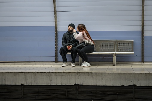 Barcelona, Spain – January 05, 2023: A young couple sitting on the bench of the Barcelona metro station, the boy dressed in black with tattoos on his face walks by