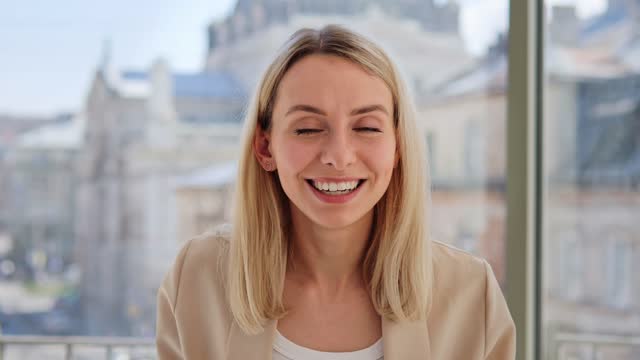 Smiling young blond woman, blogger, vlogger, influencer sitting at restaurant near a window with a blurred city view. Girl speaking looking at camera, talking making video conference call webcam view.