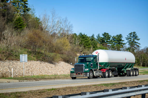 Industrial day cab green big rig semi truck transporting flammable chemicals in a specialized durable tank semi trailer driving on the two lines highway road with rocks and green forest on the side cabina diurna verde oscuro semirremolque de plataforma grande que transporta productos químicos inflamables en un semirremolque de tanque duradero especializado que conduce en la carretera de dos líneas con rocas y bosque verde en el costado