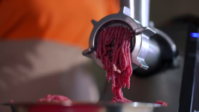 Close-up meat grinder with ground meat falling out of equipment in restaurant kitchen. Unrecognizable female Caucasian cook preparing raw ingredient for cooking indoors.