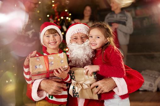 Santa Claus with Christmas presents embracing small kids at home and looking at camera.