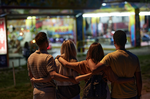 Rear view of group of friends embracing while being in the amusement park by night.