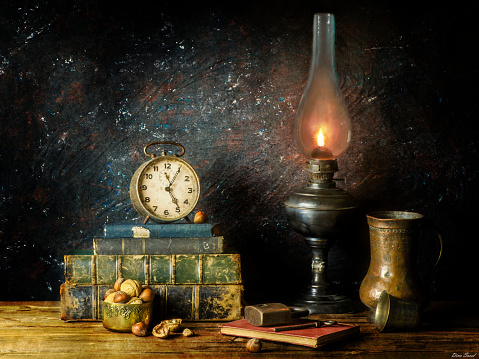 Classic still life with vintage illuminated lamp placed with old books,clock,nuts, copper jar,old key, and lock on rustic wooden background..