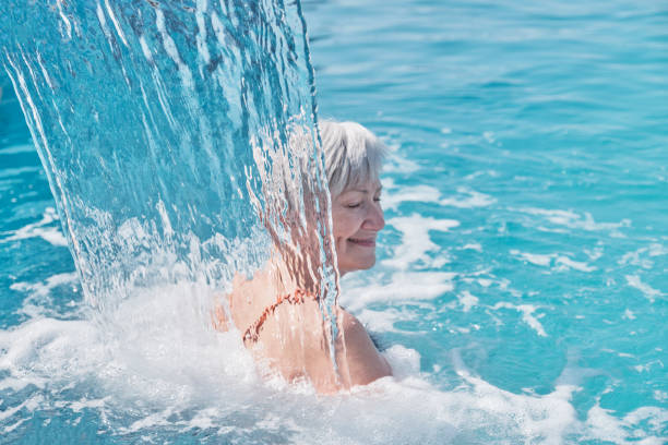 donna sorridente caucasica anziana con i capelli grigi che gode cadendo sulle sue spalle flusso d'acqua. - physical therapy hydrotherapy swimming healthcare and medicine foto e immagini stock