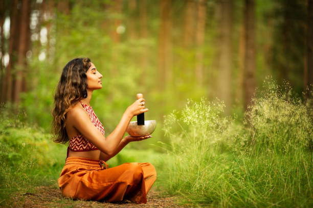 Indian Woman playing Tibetan Singing Bowls with Mallet Outdoors. Relaxing Meditative Music Therapy and Sound Healing. Spiritual Yoga Meditation Practice in Forest stock photo