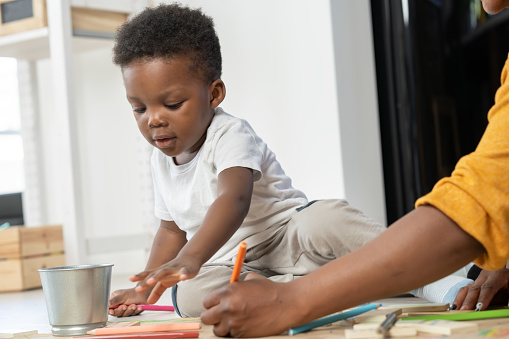 Cheerful African-American mother and cute toddler son playing with colorful pencils, drawing on a big paper, sitting on a floor. Mother-son fun bonding time. Daycare creative activities.