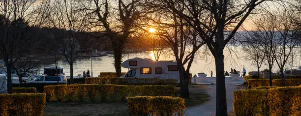 An idyllic scene from a campsite on the Adriatic coast of Croatia. The sun is setting, casting a warm glow over the beach, where children and adults are enjoying the evening. Caravans and motorhomes are seen in the backdrop.
