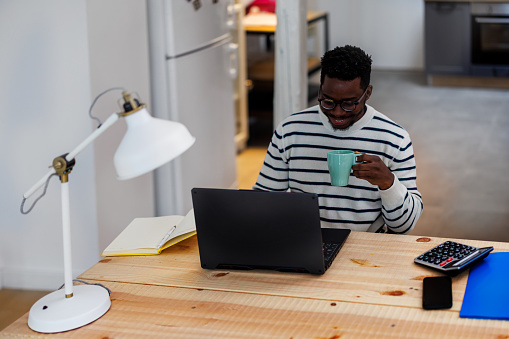 A handsome young African man is working from home using a laptop and drinking a hot cup of coffee.