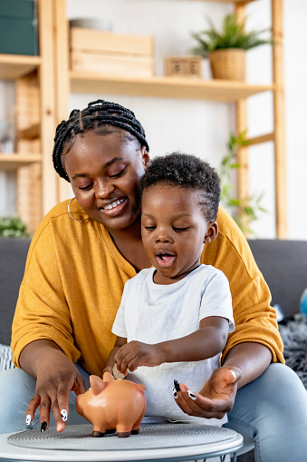 Cute little African-American boy with his mother inserting a coin into a piggy bank, financial and banking concept. Child saving money for the future concept.