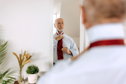 Businessman dressing up at home, tying a tie while looking at mirror
