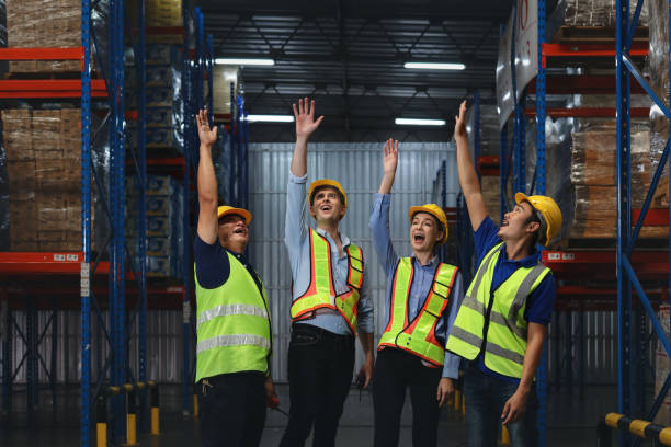 istock Group of warehouse workers with hardhats and reflective jackets standing and raising hands celebrate successful together or completed deal commitment at retail warehouse logistics, distribution center 1492837062