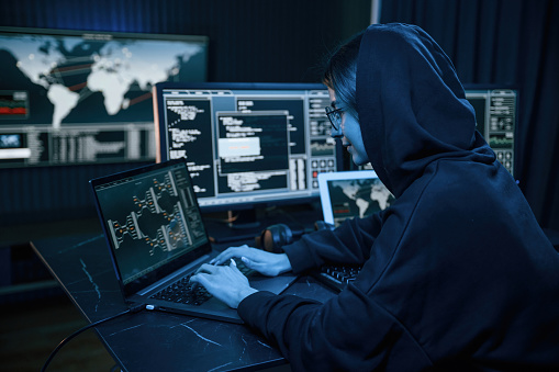 Black colored hoodie. Young professional female hacker is indoors by computer with lot of information on displays.