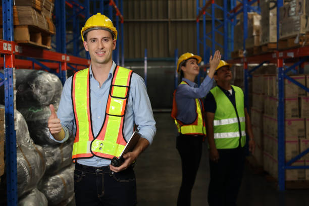 istock Group of warehouse workers with hardhats and reflective jackets using tablet, walkie talkie radio and cardboard while showing thumb up and smiling in retail warehouse logistics, distribution center 1492837479