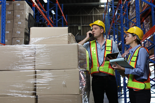 Group of warehouse workers with hardhats and reflective jackets using tablet, walkie talkie radio and cardboard while controlling stock and inventory in retail warehouse logistics, distribution center