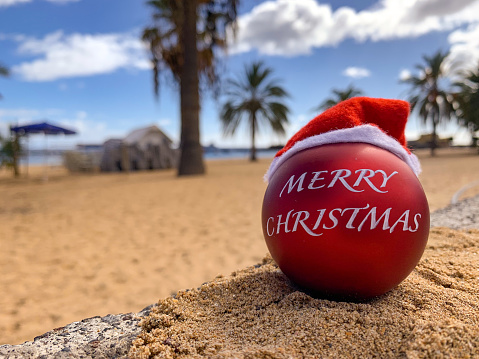 Christmas bomb in Santa's hat with words Merry Christmas in spanish on the beach lying on the sand with palm trees and blue sky on the background.