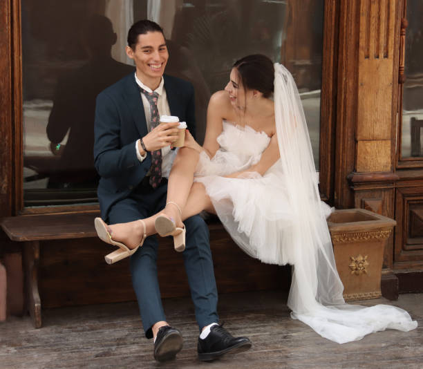 Bride and groom are drinking coffee in paper cups while sitting on bench under shop window in city. Bride and groom are having happy moment