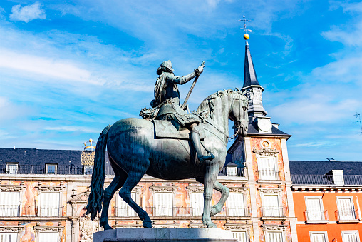 The bronze statue represents Felipe II mounted on horseback. It is placed in the centre of Plaza Mayor in Madrid and it is here represented with the clock tower as a backdrop on a partially cloudy sky during daytime.