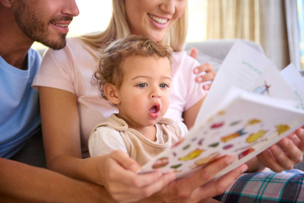 Famiglia che si siede sul divano a casa con i genitori che leggono il libro con il figlio piccolo - foto stock