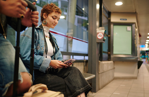 Young woman checking her mobile phone while sitting on a bench at a bus station in the early evening