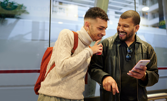 Smiling young gay couple with luggage laughing at something on a mobile phone while waiting at a bus stop